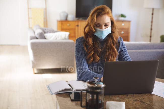 Caucasian woman spending time at home, in the kitchen, working from home, using her laptop,  wearing a face mask. Social distancing during Covid 19 Coronavirus quarantine lockdown. — Stock Photo