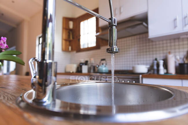 Primo piano di un rubinetto da cucina con acqua che scorre in un lavandino in acciaio in una cucina moderna con armadi sfocati sullo sfondo. Interni design idea cucina. — Foto stock