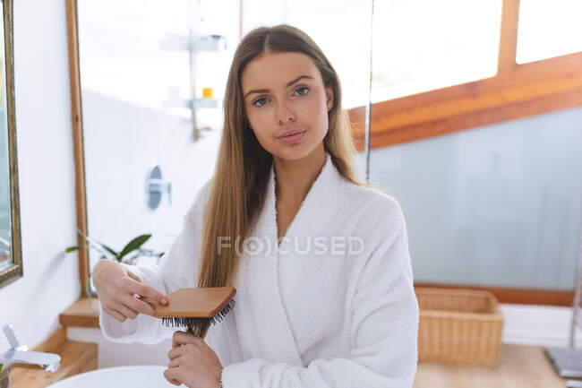 Retrato de mulher caucasiana passando tempo em casa, de pé no banheiro, olhando para a câmera escovando o cabelo olhando para a câmera. Distanciamento social durante o bloqueio de quarentena do Covid 19 Coronavirus. — Fotografia de Stock