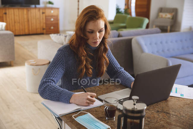 Mujer caucásica pasando tiempo en casa, en la cocina, trabajando desde casa, usando su computadora portátil y escribiendo. Distanciamiento social durante el bloqueo de cuarentena del Coronavirus Covid 19. - foto de stock