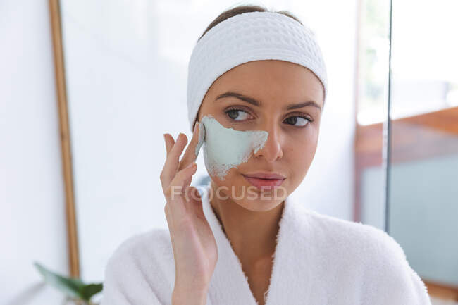 Mujer caucásica pasar tiempo en casa, de pie en el baño, mirando en el espejo aplicando mascarilla facial. Distanciamiento social durante el bloqueo de cuarentena del Coronavirus Covid 19. - foto de stock