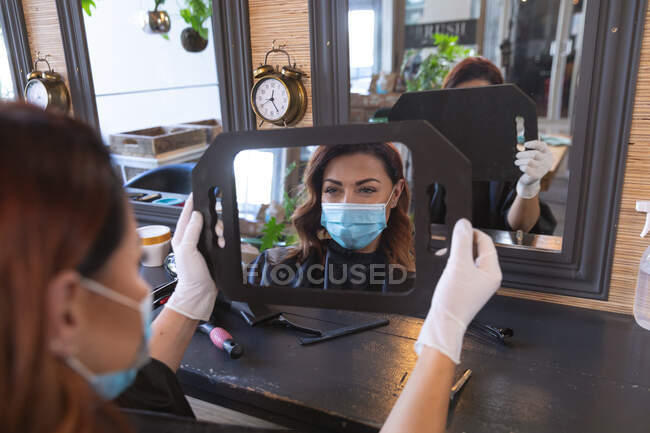 Белая женщина-клиент парикмахера в маске и смотрит на свое отражение в зеркале. Здоровье и гиперактивность на рабочем месте во время коронавируса Ковид 19 пандемии. — стоковое фото