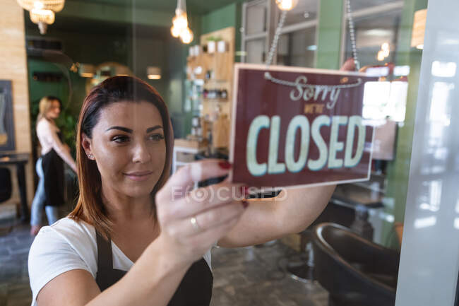 Caucasian female hairdresser working in hair salon smiling, fixing a sign saying Sorry, We Are Closed. Health and hygiene in workplace during Coronavirus Covid 19 pandemic. — Stock Photo