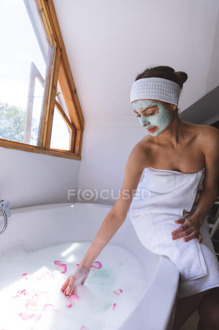 Femme caucasienne passant du temps à la maison, dans la salle de bain avec masque facial, jetant des pétales dans l'eau, assise sur le bord de la baignoire. Distance sociale pendant le confinement en quarantaine du coronavirus Covid 19. — Photo de stock