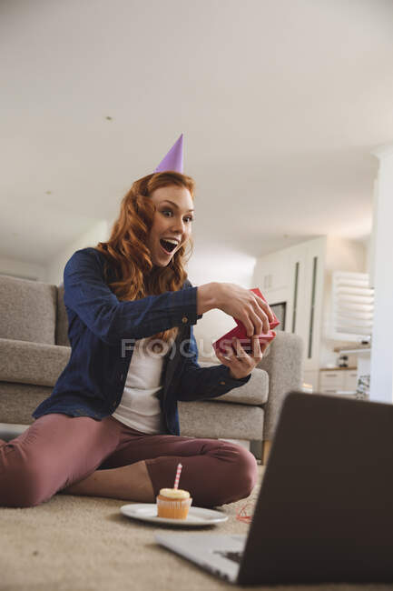 Mujer caucásica pasando tiempo en casa, en la sala de estar, sonriendo, celebrando, abriendo un regalo, magdalena a su lado. Distanciamiento social durante el bloqueo de cuarentena del Coronavirus Covid 19. — Stock Photo