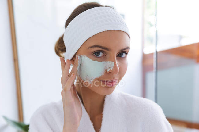 Портрет белой женщины, проводящей время дома, стоящей в ванной, смотрящей в зеркало в маску. Социальное дистанцирование во время изоляции коронавируса Covid 19. — стоковое фото
