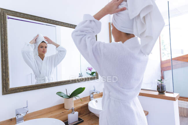 Kaukasische Frau verbringt Zeit zu Hause, steht im Badezimmer und schaut in Spiegel, der Handtuch um ihr Haar wickelt. Soziale Distanzierung während Covid 19 Coronavirus Quarantäne Lockdown. — Stockfoto