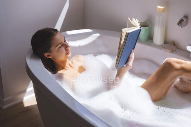 Donna caucasica trascorrere del tempo a casa, in bagno, sdraiato nella vasca da bagno, libro di lettura. Distanza sociale durante il blocco di quarantena Covid 19 Coronavirus. — Foto stock