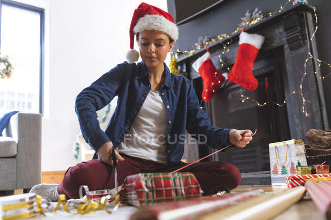 Femme caucasienne passant du temps à la maison à Noël, portant le chapeau de Père Noël, assise sur le sol dans le salon, enveloppant présent dans le papier. Distance sociale pendant le confinement en quarantaine du coronavirus Covid 19. — Photo de stock