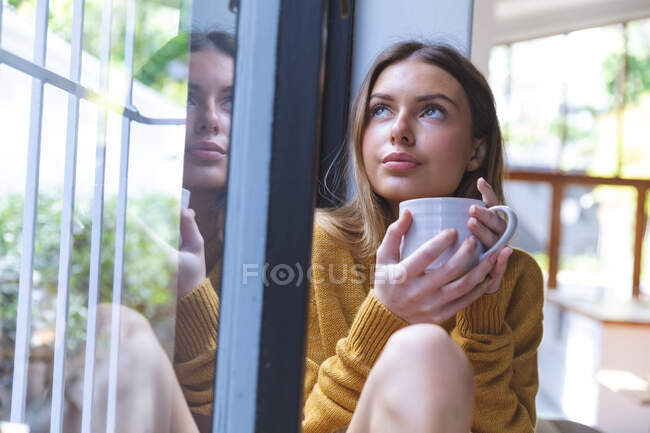 Femme caucasienne passant du temps à la maison, assise par la fenêtre, tenant une tasse verte regardant par la fenêtre. Distance sociale pendant le confinement en quarantaine du coronavirus Covid 19. — Photo de stock