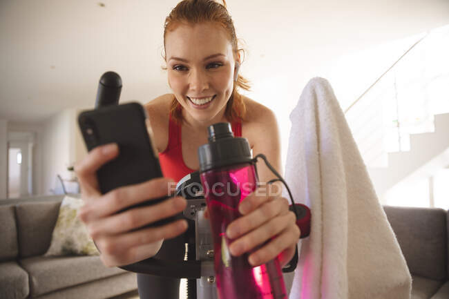 Mulher caucasiana passando tempo em casa, na sala de estar, exercitando-se em bicicleta estacionária, usando seu smartphone. Distanciamento social durante o bloqueio de quarentena do Covid 19 Coronavirus. — Fotografia de Stock