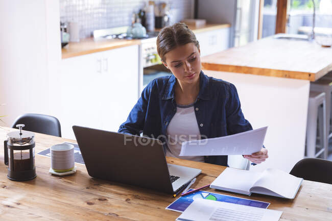 Mujer caucásica pasar tiempo en casa, sentado junto a la mesa en la cocina utilizando el ordenador portátil, trabajando desde casa. Distanciamiento social durante el bloqueo de cuarentena del Coronavirus Covid 19. - foto de stock