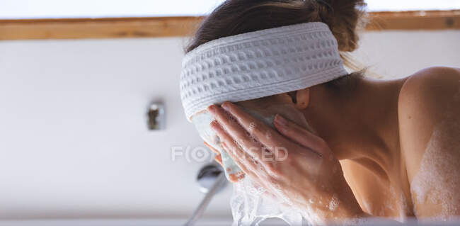 Белая женщина проводит время дома, в ванной, носит повязку на голове, смывает маску с лица. Социальное дистанцирование во время изоляции коронавируса Covid 19. — стоковое фото