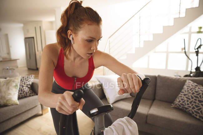 Белая женщина проводит время дома, в гостиной, тренируется на стационарном велосипеде, проверяет свои умные часы. Социальное дистанцирование во время изоляции коронавируса Covid 19. — стоковое фото