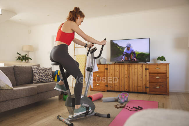 Femme caucasienne passant du temps à la maison, dans le salon, faisant de l'exercice à vélo stationnaire, regardant la télévision. Distance sociale pendant le confinement en quarantaine du coronavirus Covid 19. — Photo de stock