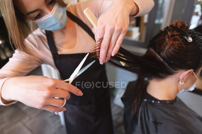 Caucasian female hairdresser working in hair salon wearing face mask, cutting hair of female Caucasian customer in face mask. Health and hygiene in workplace during Coronavirus Covid 19 pandemic. — Stock Photo