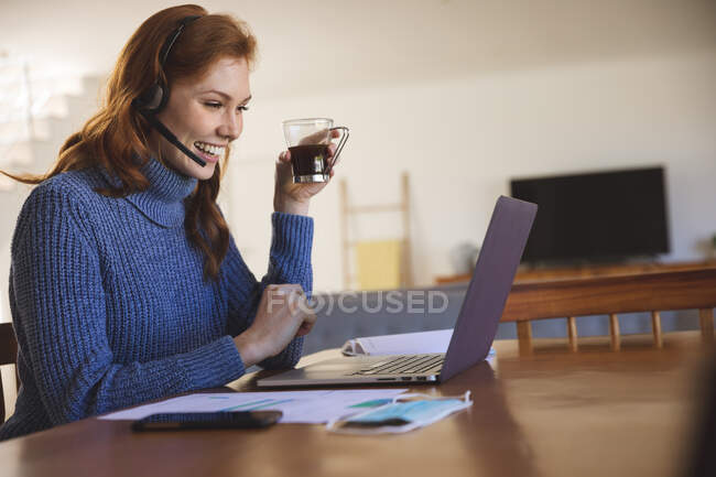 Caucasian woman spending time at home, in the kitchen, working from home, using her laptop, wearing a headset, smiling. Social distancing during Covid 19 Coronavirus quarantine lockdown. — Stock Photo