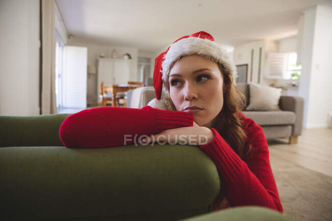 Кавказька жінка проводить час удома, у вітальні, виглядаючи сумною, одягнувши різдвяний капелюх і червоний стрибун. Соціальна дистанція в Ковиді 19 Коронавірус карантин. — стокове фото