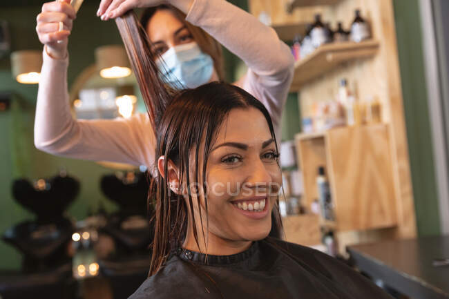 Кавказька жіноча перукарка, що працює в салоні для волосся, одягнена в маску обличчя, стрижка волосся жінки-кавказької клієнтки. Здоров 
