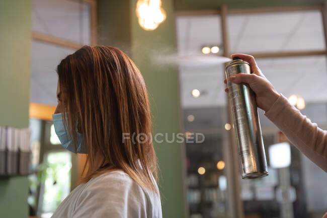 Caucasian female hairdresser working in hair salon spraying hairspray on hair of female Caucasian customer in face mask. Health and hygiene in workplace during Coronavirus Covid 19 pandemic. — Stock Photo