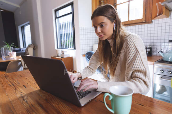 Caucasian woman spending time at home, sitting in kitchen using laptop computer with earphones on. Social distancing during Covid 19 Coronavirus quarantine lockdown. — Stock Photo