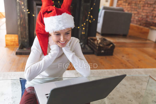 Femme caucasienne passe du temps à la maison à Noël, portant le chapeau de Père Noël, assis à table à l'aide d'un ordinateur pendant le chat vidéo. Distance sociale pendant le confinement en quarantaine du coronavirus Covid 19. — Photo de stock