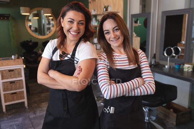 Portrait de deux coiffeuses caucasiennes travaillant dans un salon de coiffure, posant pour une photo avec les bras croisés. Santé et hygiène sur le lieu de travail pendant la pandémie de Coronavirus Covid 19. — Photo de stock