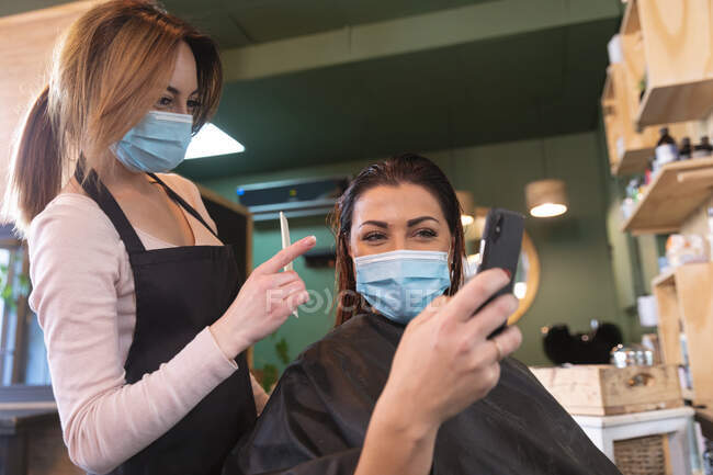 Kaukasische Friseurin, die im Friseursalon arbeitet, trägt Gesichtsmaske, kämmt die Haare einer kaukasischen Kundin in Gesichtsmaske. Gesundheit und Hygiene am Arbeitsplatz während der Coronavirus Covid 19 Pandemie. — Stockfoto