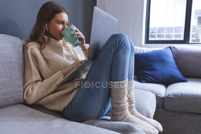 Кавказка проводит время дома, сидит на диване в гостиной, используя ноутбук с наушниками, держит кружку, пьет. Социальное дистанцирование во время изоляции коронавируса Covid 19. — стоковое фото
