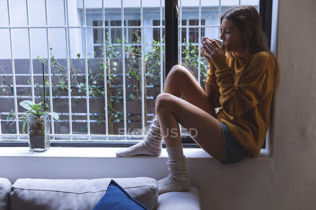 Caucasian woman spending time at home, sitting on windowsill in living room, drinking from green mug. Social distancing during Covid 19 Coronavirus quarantine lockdown. — Stock Photo