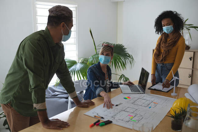 Multi ethnic group of male and female architects in office wearing face masks, discussing over architectural drawing. Health and hygiene in workplace during Coronavirus Covid 19 pandemic. — Stock Photo