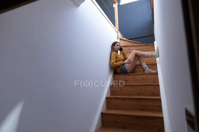 Femme caucasienne passant du temps à la maison, assise dans les escaliers dans le couloir, parlant sur son smartphone. Distance sociale pendant le confinement en quarantaine du coronavirus Covid 19. — Photo de stock