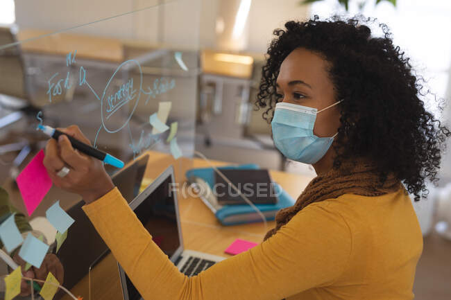 Mista raça fêmea criativa em máscara facial trabalhando na mesa no escritório, escrevendo na tela protetora. Saúde e higiene no local de trabalho durante a pandemia de Coronavirus Covid 19. — Fotografia de Stock