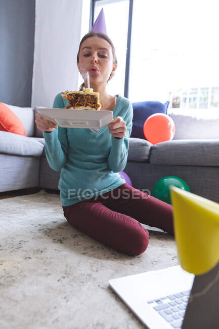 Счастливая белая женщина проводит время дома, в шляпе, сидит на полу, используя компьютер во время видео чата, задувая свечу. Социальное дистанцирование во время изоляции коронавируса Covid 19. — стоковое фото