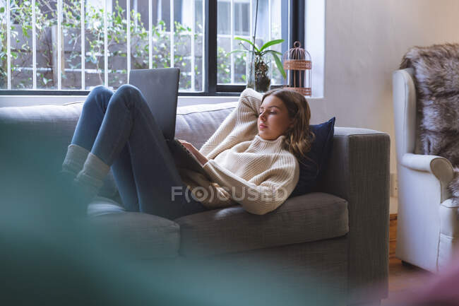 Mujer caucásica pasar tiempo en casa, acostado en el sofá en la sala de estar con ordenador portátil. Distanciamiento social durante el bloqueo de cuarentena del Coronavirus Covid 19. - foto de stock
