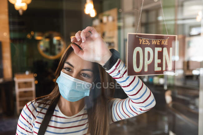 Cabeleireiro feminino caucasiano trabalhando no salão de cabeleireiro usando máscara facial, apoiando-se na porta de vidro com um sinal dizendo que está aberto. Saúde e higiene no local de trabalho durante a pandemia de Coronavirus Covid 19. — Fotografia de Stock