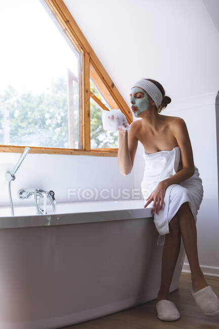Белая женщина проводит время дома, в ванной комнате в маске для лица, сидя на краю ванны, сдувая пену с руки. Социальное дистанцирование во время изоляции коронавируса Covid 19. — стоковое фото