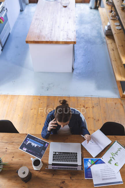 Белая женщина проводит время дома, сидит за столом с ноутбуком, работает из дома, разговаривает на смартфоне. Социальное дистанцирование во время изоляции коронавируса Covid 19. — стоковое фото