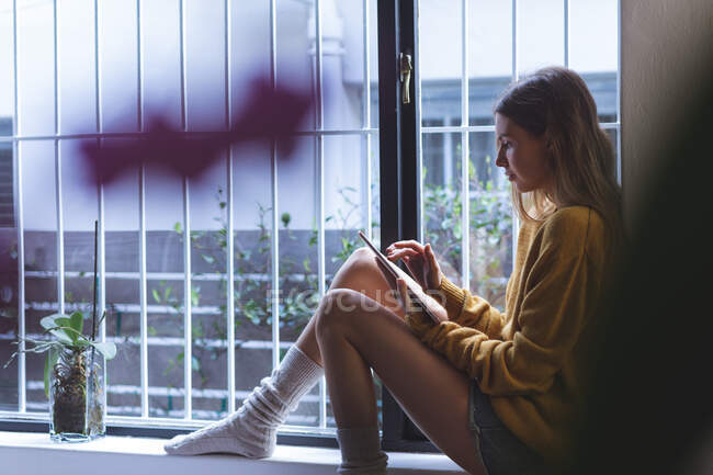 Caucasian woman spending time at home, sitting on windowsill in living room, holding digital tablet. Social distancing during Covid 19 Coronavirus quarantine lockdown. — Stock Photo