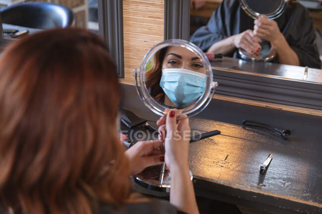 Caucasian female customer of a hairdresser wearing a face mask and looking at her reflection in the mirror. Health and hygiene in workplace during Coronavirus Covid 19 pandemic. — Stock Photo