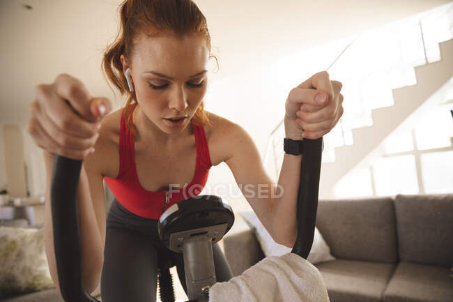 Femme caucasienne passant du temps à la maison, dans le salon, faisant de l'exercice sur un vélo fixe avec ses écouteurs dedans. Distance sociale pendant le confinement en quarantaine du coronavirus Covid 19. — Photo de stock
