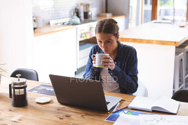 Caucasian woman spending time at home, sitting by table in kitchen using laptop computer, working from home, drinking from mug. Social distancing during Covid 19 Coronavirus quarantine lockdown. — Stock Photo