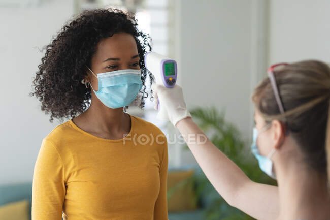 Caucasian woman using digital thermometer taking temperature of mixed race woman arriving at work wearing face mask. Health and hygiene in workplace during Coronavirus Covid 19 pandemic. — Stock Photo