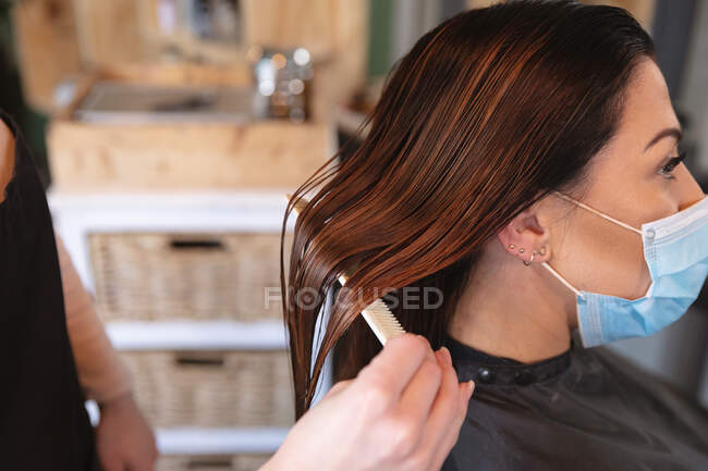 Белая женщина-парикмахер работает в парикмахерской, расчесывая волосы белой клиентки в маске. Здоровье и гиперактивность на рабочем месте во время коронавируса Ковид 19 пандемии. — стоковое фото