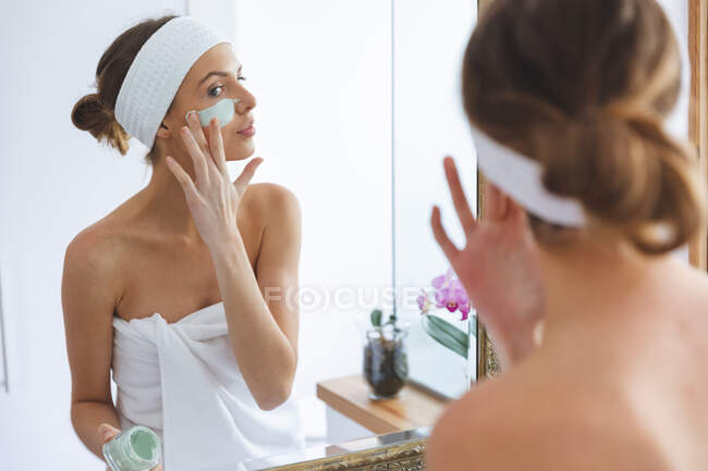 Кавказька жінка проводить час удома, стоїть у ванній кімнаті і дивиться у дзеркало, застосовуючи маску обличчя. Соціальна дистанція в Ковиді 19 Коронавірус карантин. — стокове фото