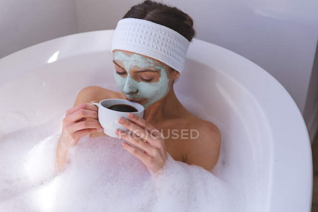 Femme caucasienne passant du temps à la maison, dans la salle de bain avec masque, assise dans la baignoire, buvant du café. Distance sociale pendant le confinement en quarantaine du coronavirus Covid 19. — Photo de stock