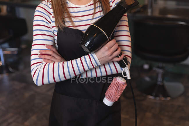 Mid section of female hairdresser working in hair salon, holding a hair dryer with her arms crossed. Health and hygiene in workplace during Coronavirus Covid 19 pandemic. — Stock Photo