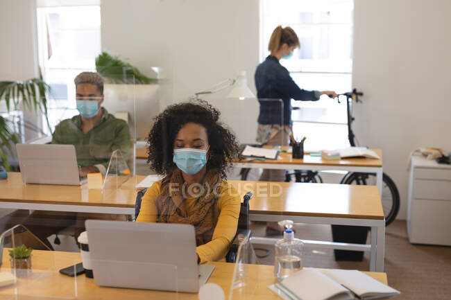 Grupo multiétnico de creativos masculinos y femeninos que trabajan en escritorios de oficina con pantallas protectoras, utilizando computadoras portátiles. Salud e higiene en el lugar de trabajo durante la pandemia de Coronavirus Covid 19. - foto de stock