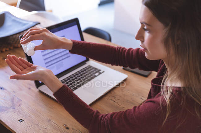 Caucasian woman spending time at home, sitting by table with laptop on, sanitizing her hands. Social distancing during Covid 19 Coronavirus quarantine lockdown. — Stock Photo