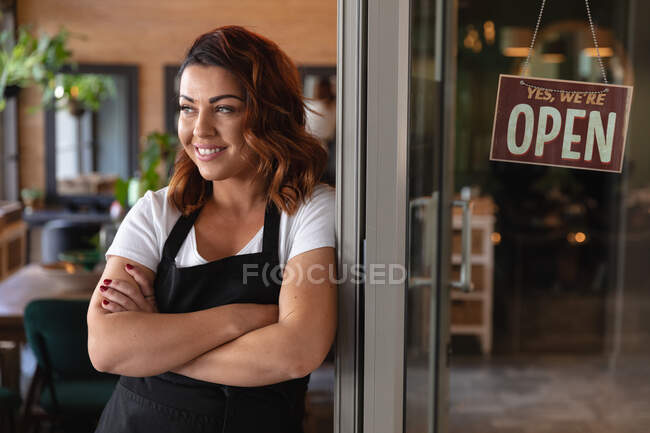 Caucasian female hairdresser working in hair salon smiling, leaning on glass door with a sign saying Yes, We Are Open. Health and hygiene in workplace during Coronavirus Covid 19 pandemic. — Stock Photo